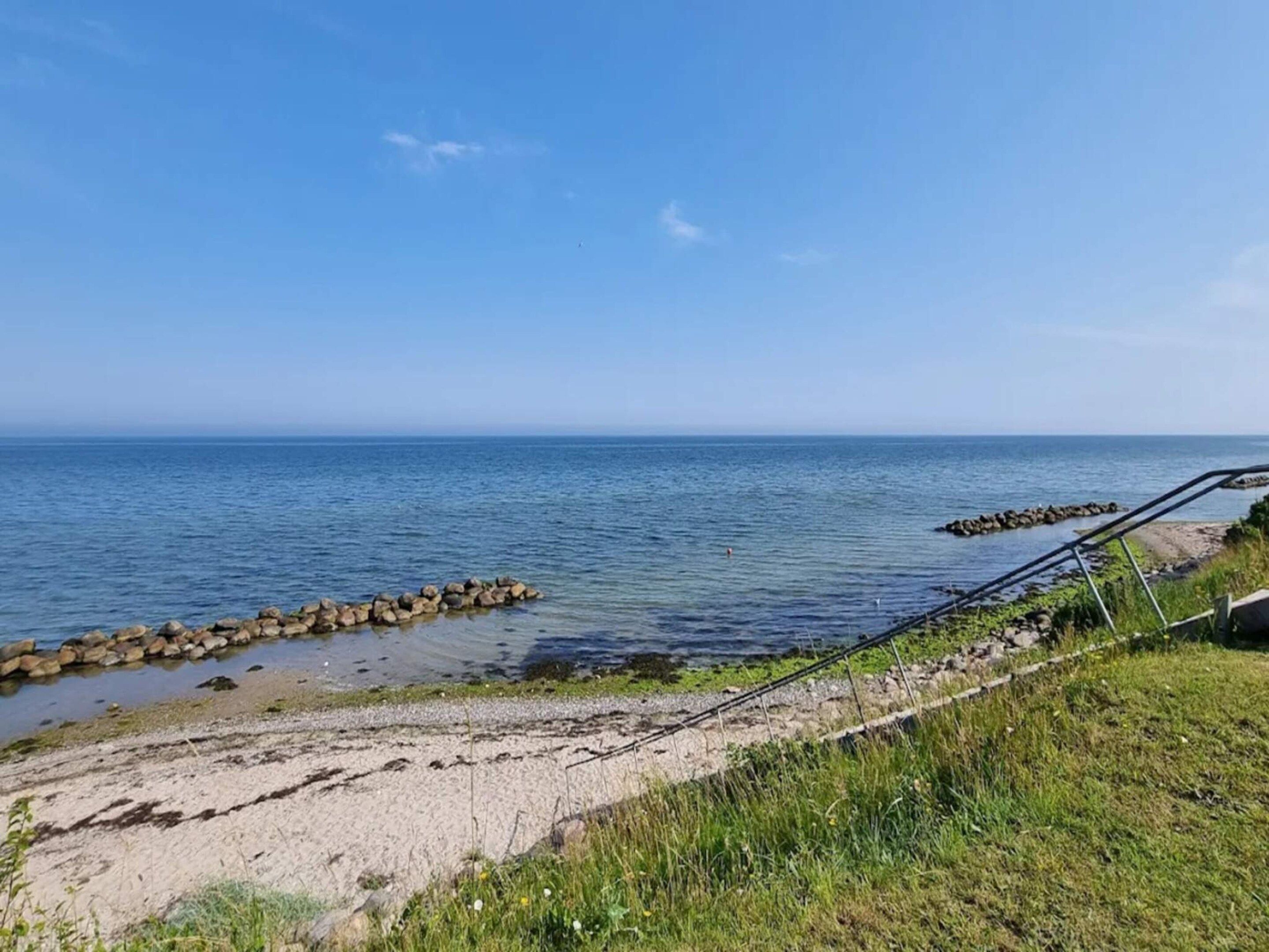 A coastal view with a sandy beach, a rocky breakwater, and a clear blue sky.
