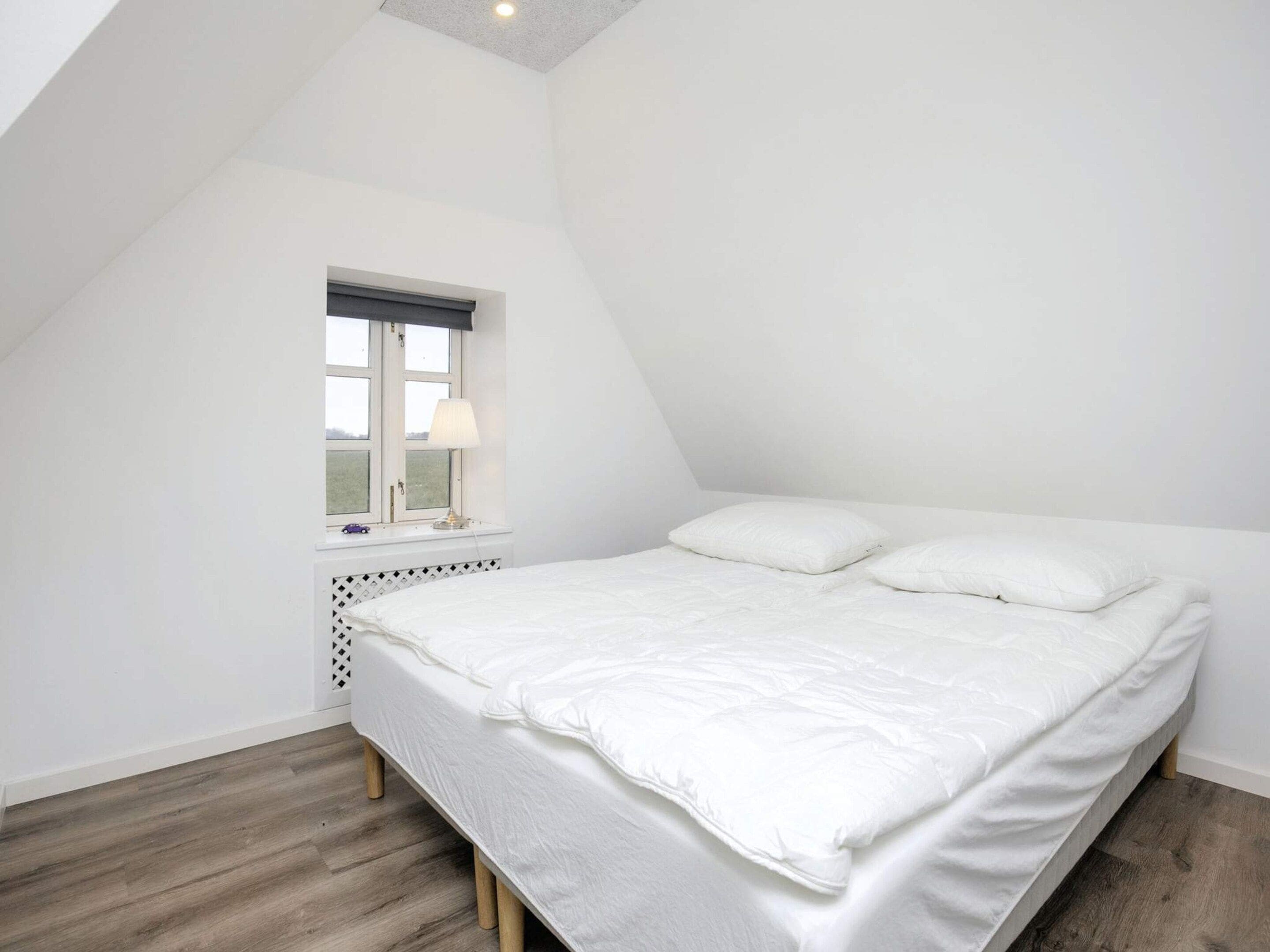 Un dormitorio con techo inclinado, una ventana con lámpara, una cama con almohadas y una mesita con lámpara.