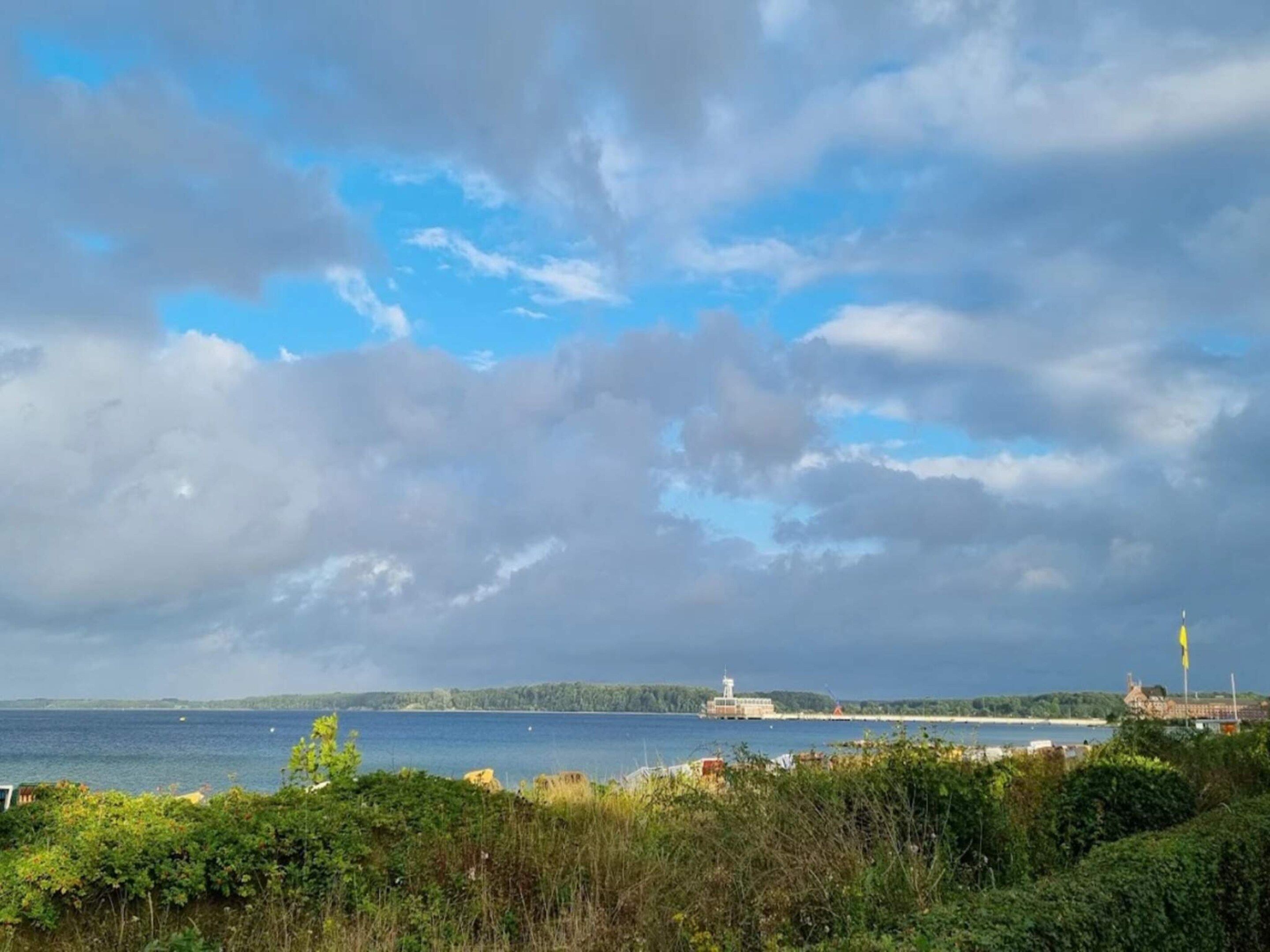 A coastal landscape with a lighthouse, dense greenery, and a body of water under a partly cloudy sky.