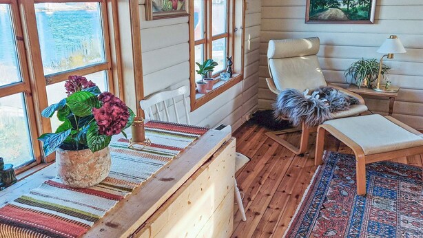 A cozy cabin interior with wooden walls, a large window, a comfortable chair, a small table with a plant, and a rug. - 7 Person Holiday Home in Averoy (Averoy)