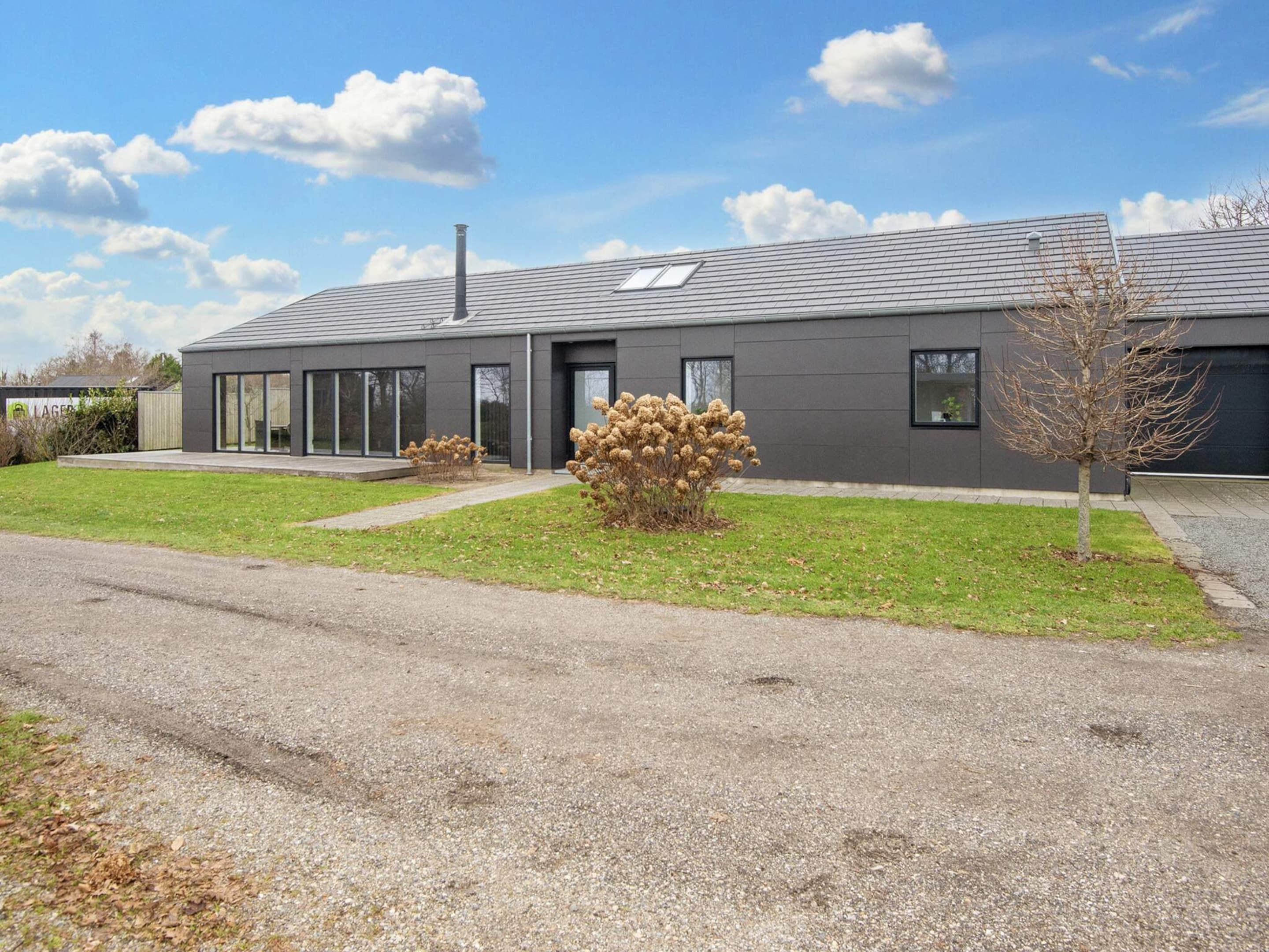 A modern two-story house with a grey roof, large windows, and a gravel driveway.