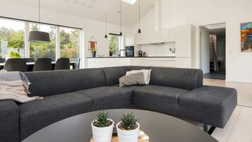 A modern living room with a grey sectional sofa, a round coffee table with potted plants, and a view of the kitchen and dining area through large windows.