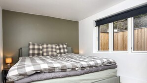 A bed with a checkered bedspread, a nightstand with a lamp, and a window with a view of a fence. - 4 Star Holiday Home in Ebeltoft (Ebeltoft)