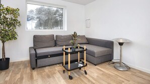 A modern living room with a grey sofa, a small black and wood side table with a plant, a floor lamp, and a window with a view of trees. - 4 Star Holiday Home in Ebeltoft (Ebeltoft)