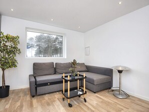 A modern living room with a grey sofa, a small black and wood side table with a plant, a floor lamp, and a window with a view of trees. - 4 Star Holiday Home in Ebeltoft (Ebeltoft)
