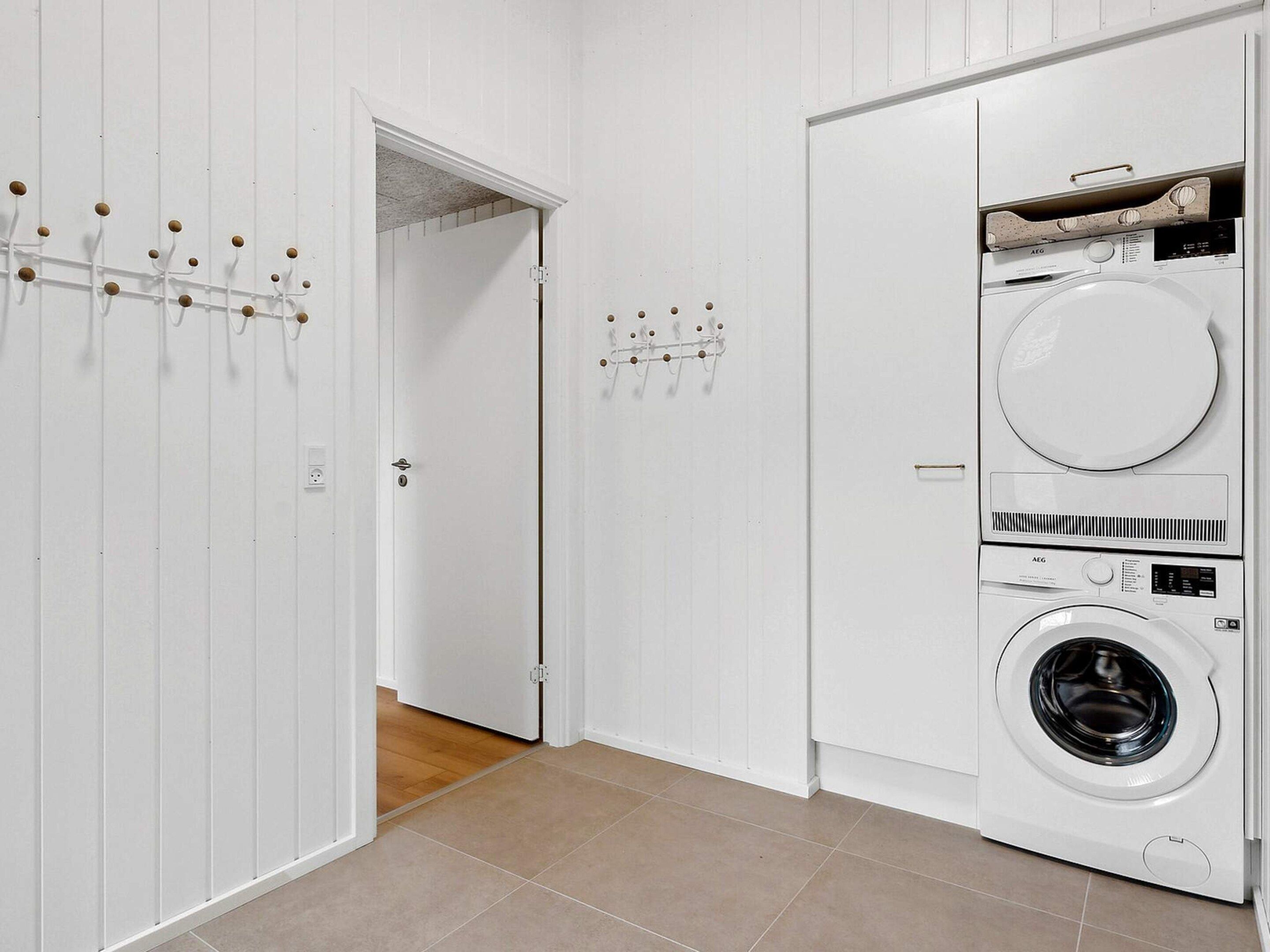 Et vaskerum med en frontbetjent vaskemaskine og tørretumbler, en dør med knagerække og et flisegulv.