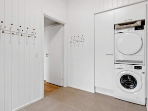 A laundry room with a front-loading washer and dryer, a door with a coat rack, and a tiled floor. - 5 Star Holiday Home in Henne (Henne)