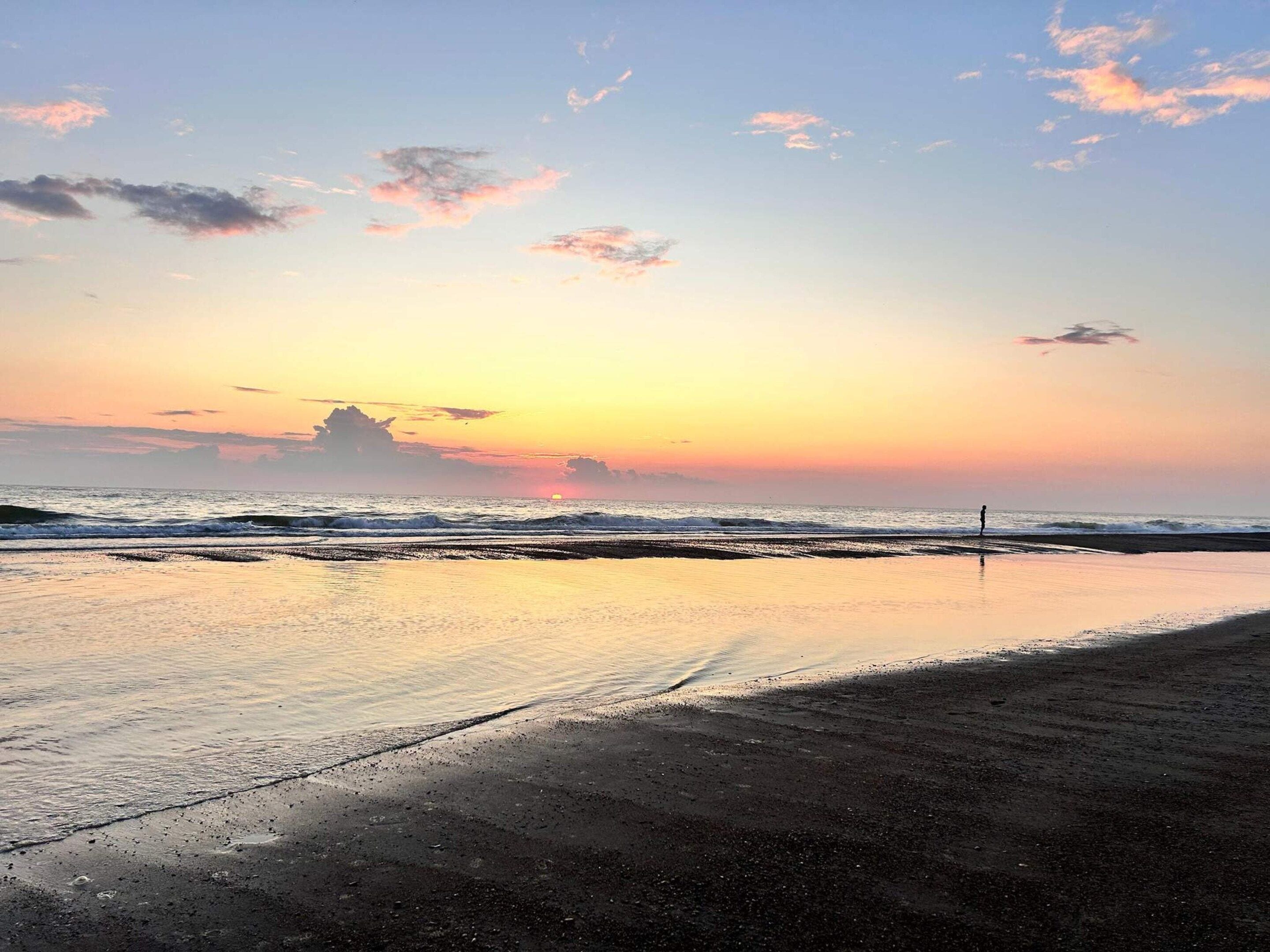 Una playa con un atardecer, olas y una persona de pie en la orilla.