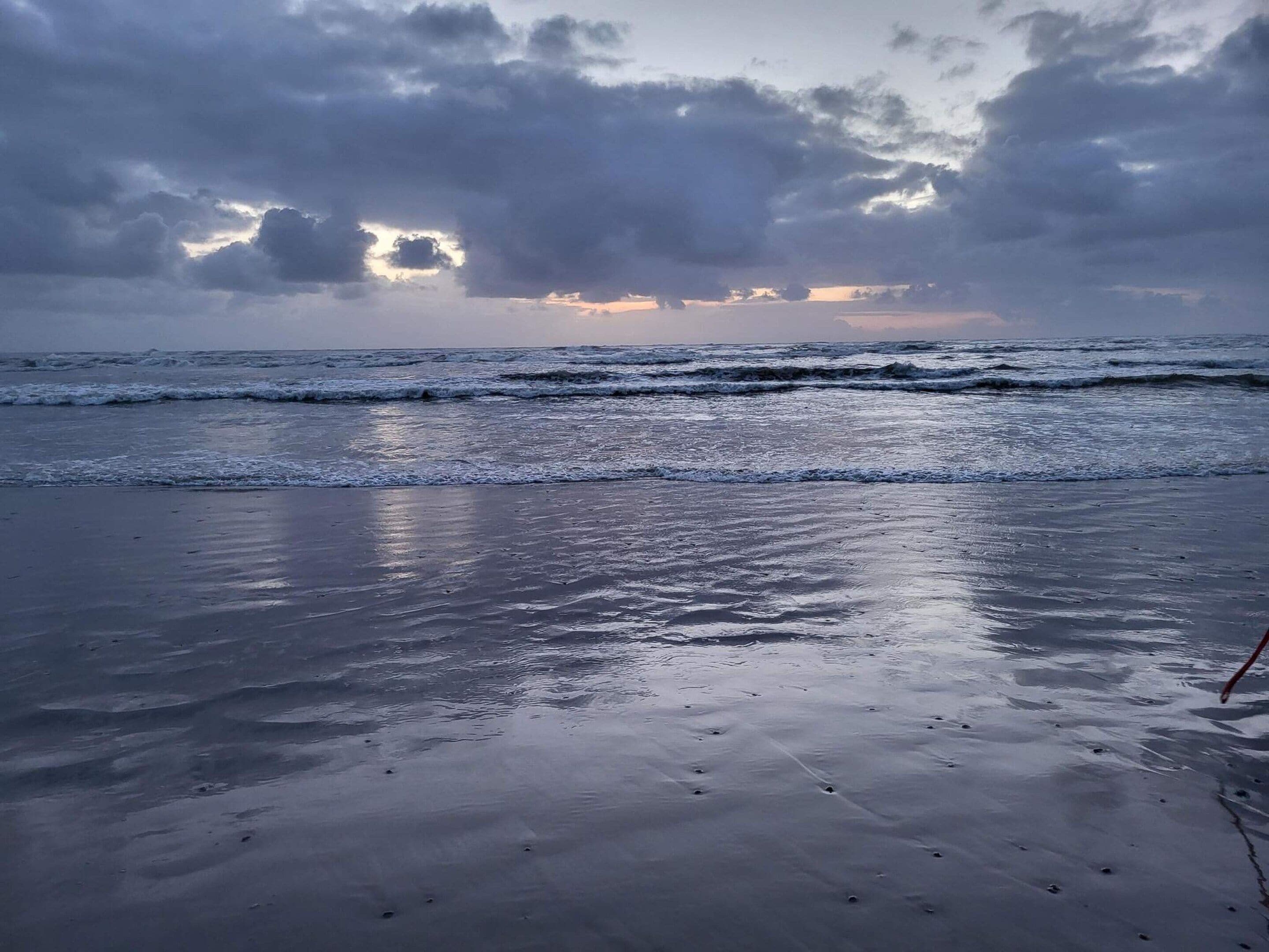 Een strand met nat zand en golven, onder een bewolkte lucht.