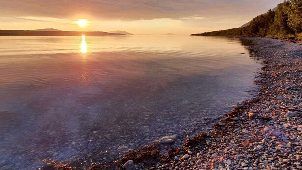 Ein ruhiger See bei Sonnenuntergang, mit kiesigem Ufer und fernen Bergen.