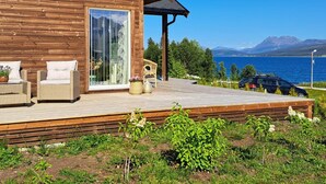 Eine Holzhütte mit Terrasse, Korbmöbeln und Blick auf einen See und Berge.