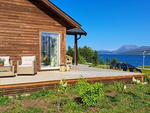 A wooden cabin with a deck, wicker furniture, and a view of a lake and mountains. - 6 Person Holiday Home in Svensby (Lyngen)