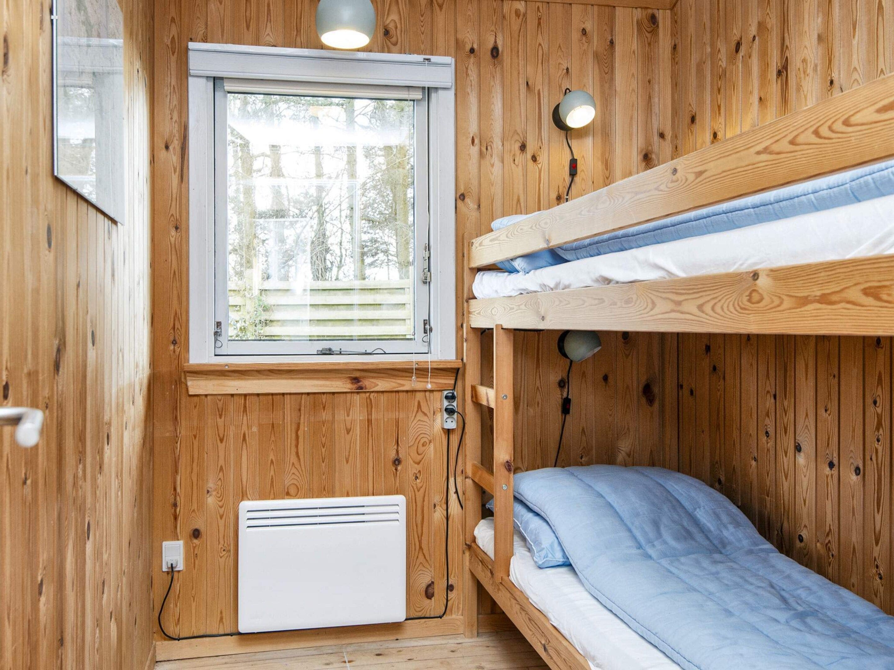 A compact room with a bunk bed, a window with blinds, a wall-mounted heater, and a small table with a lamp.