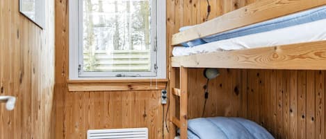 A compact room with a bunk bed, a window with blinds, a wall-mounted heater, and a small table with a lamp.