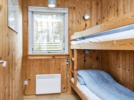 Habitación pequeña con una cama litera, ventana con persianas, calentador de pared y una pequeña mesa con lámpara.