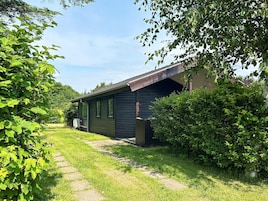 A small, single-story cabin with a sloped roof, surrounded by lush greenery and a stone pathway leading to the entrance.