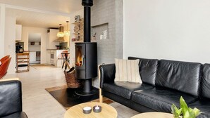 A modern living room with a black wood-burning stove, a black leather sofa, and a dining area in the background. - 8 Person Holiday Home in Oksbol (Oksbol)