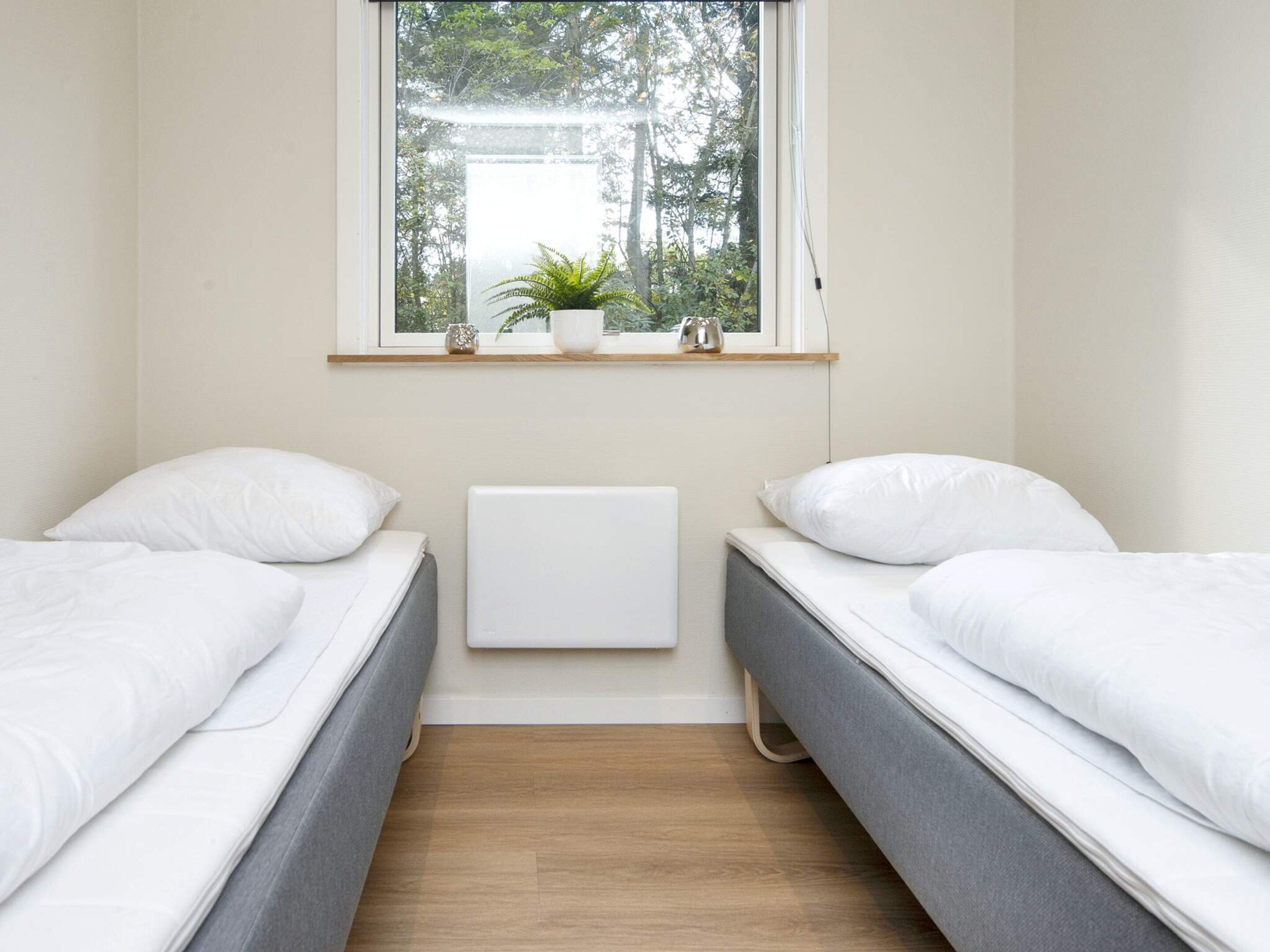 A bedroom with two single beds, a window with a view of trees, a radiator, and a shelf with a plant and a vase.