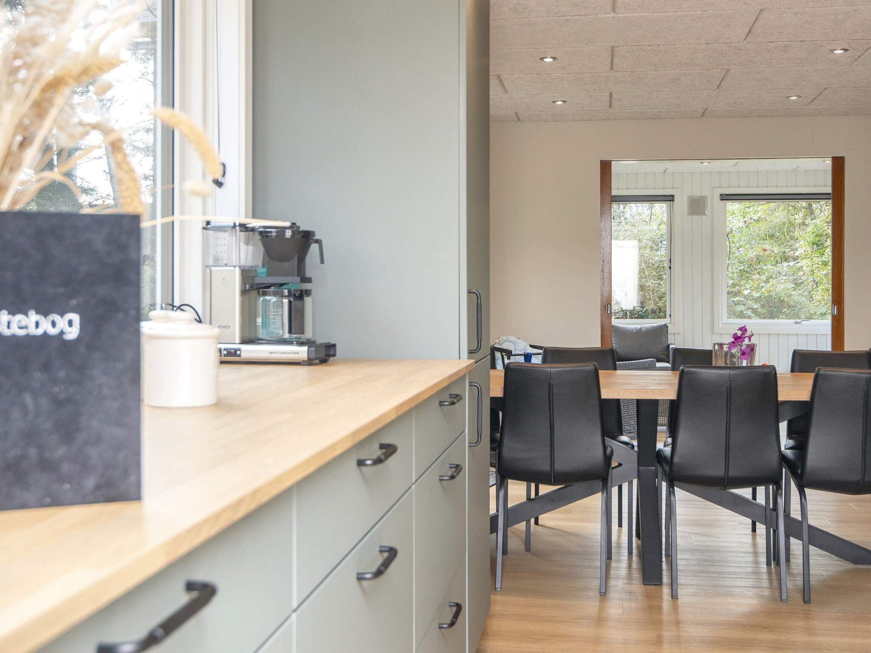 A modern kitchen with a coffee machine, a dining table with chairs, and a view of greenery outside.