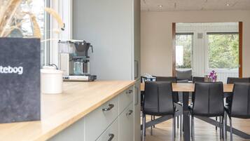 A modern kitchen with a coffee machine, a dining table with chairs, and a view of greenery outside.