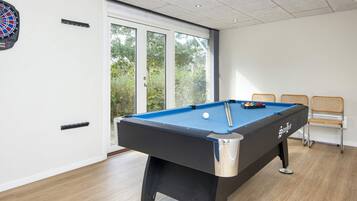 A pool table with a dartboard and chairs in a room with large windows.