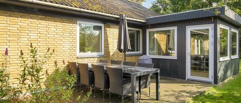 Ein modernes Haus mit einer Terrasse, einem Tisch, Stühlen, großen Fenstern und einer Holzterrasse.