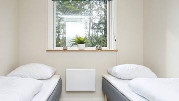 A room with two single beds, a window with a view of trees, a radiator, and a shelf with a plant and a vase.