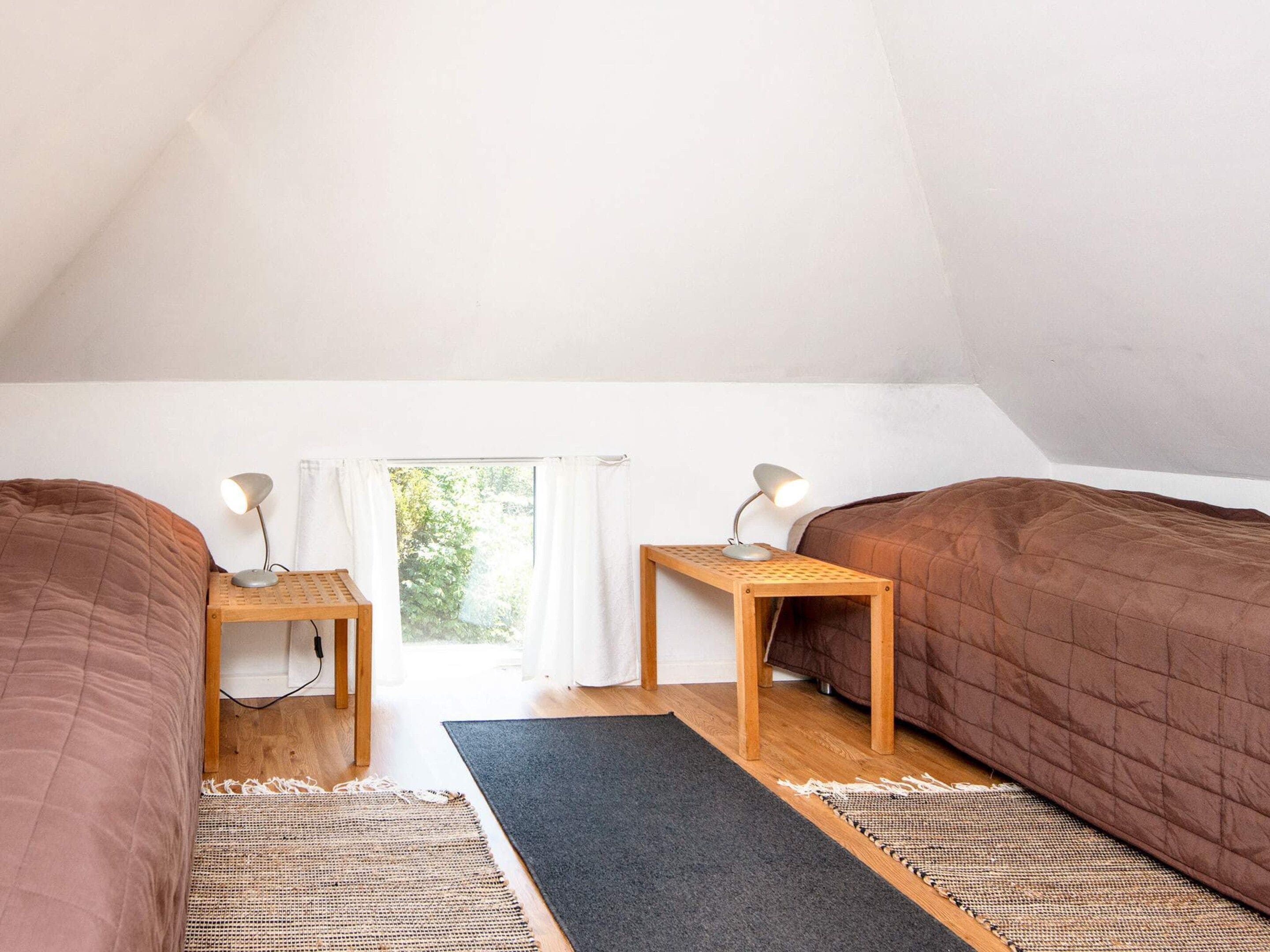 A room with two beds, a window with curtains, and a view of greenery outside.