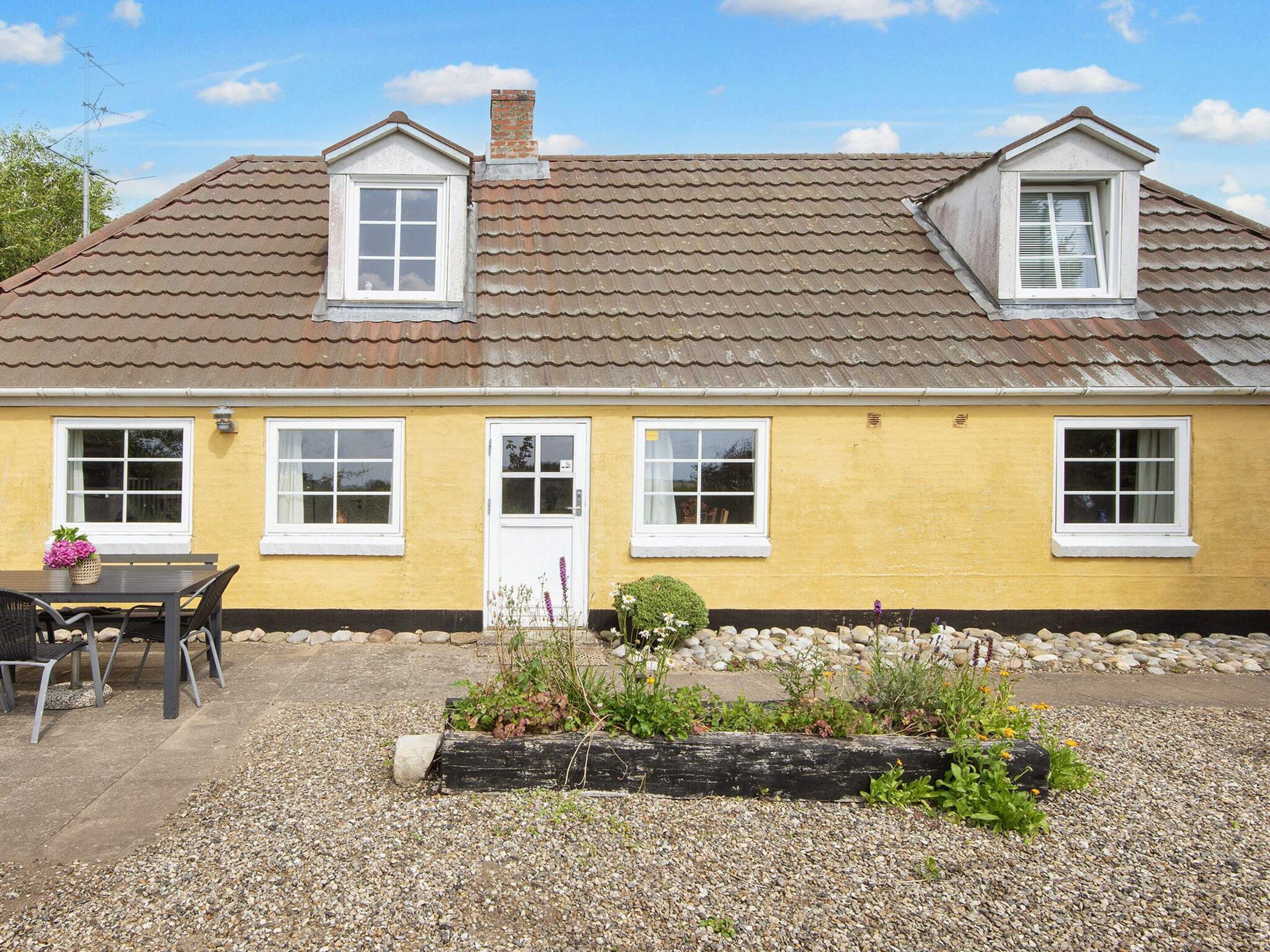 A yellow house with a tiled roof, a front door, and a small garden area with gravel and plants.