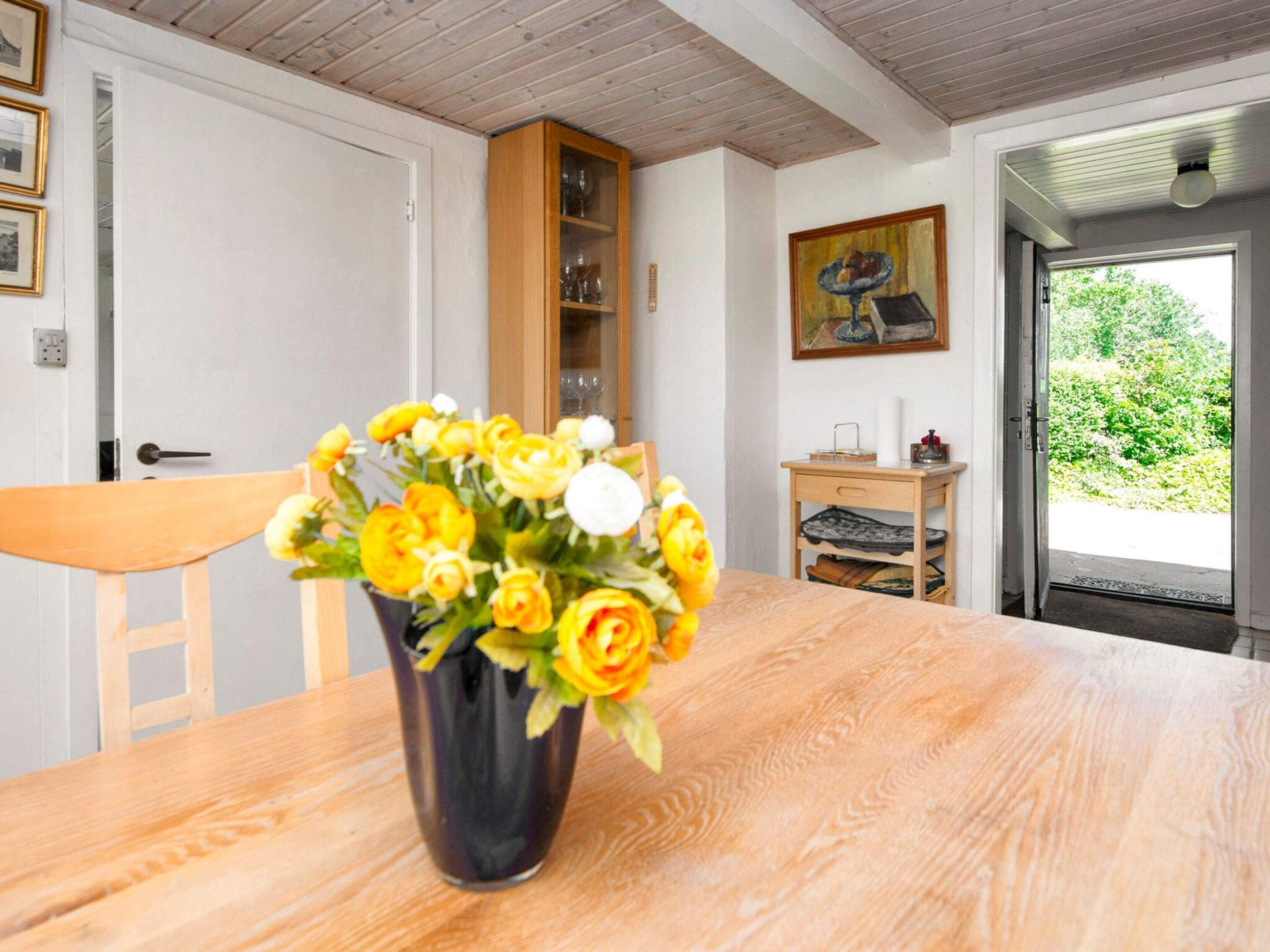 A dining table with a vase of yellow flowers, a wooden chair, a cabinet with glassware, and a painting on the wall.