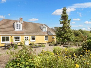 A yellow house with a tiled roof, a chimney, and a small porch with a table and chairs. - 6 Person Holiday Home in Ringkobing (Ringkobing)