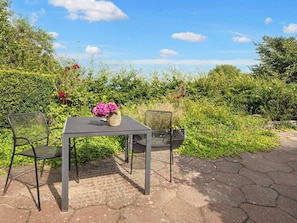 An outdoor seating area with a table and two chairs, surrounded by lush greenery and a clear sky. - 6 Person Holiday Home in Ringkobing (Ringkobing)