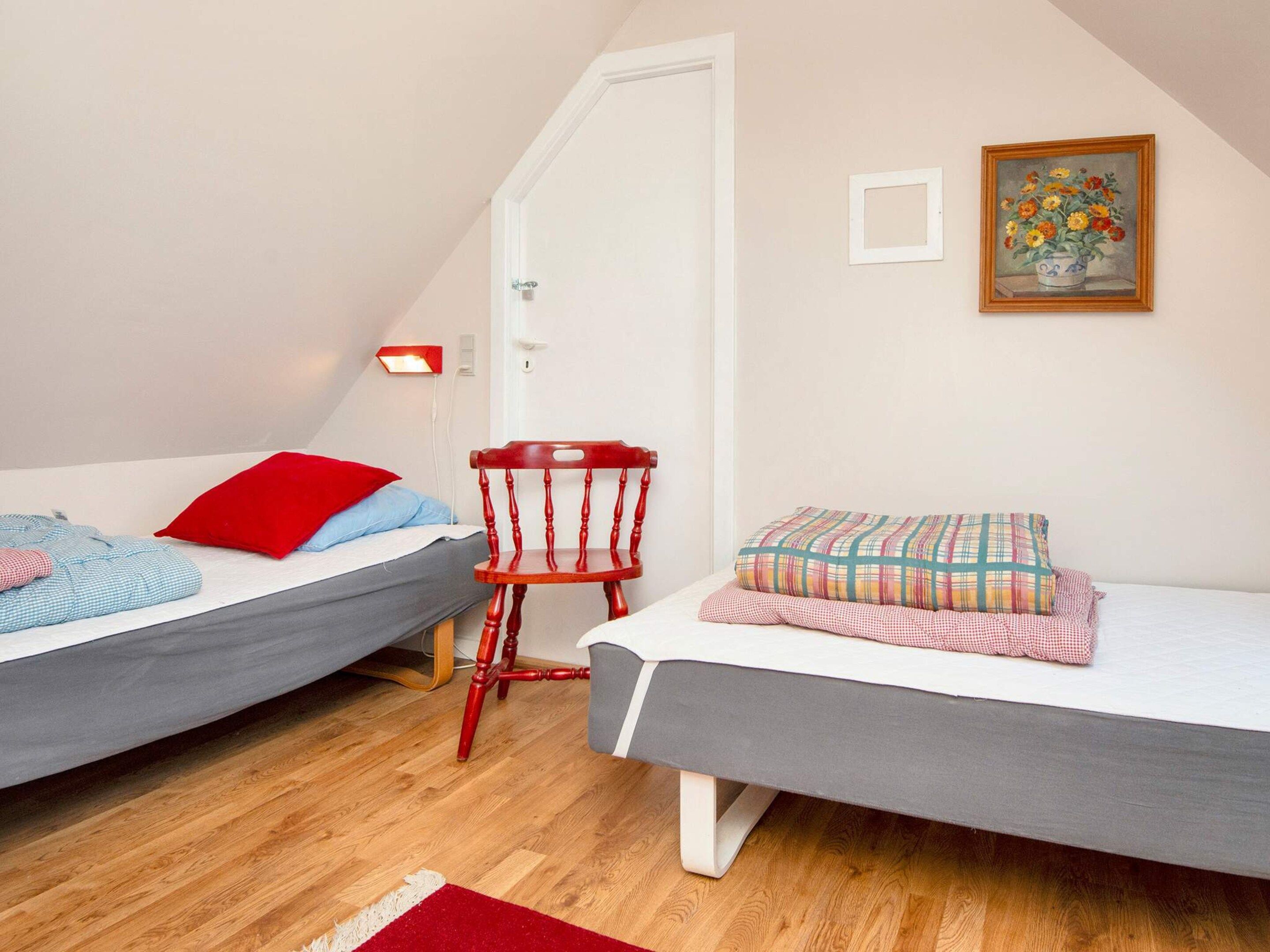 A bedroom with two single beds, a wooden chair, a red pillow, a plaid pillow, a painting, and a red rug.