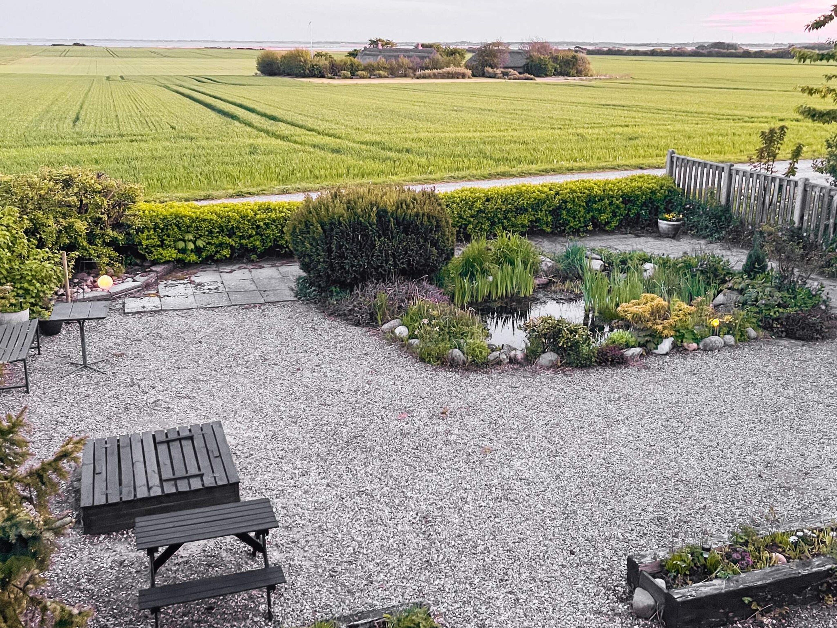 A gravel patio with a wooden bench, surrounded by a garden with various plants and a small pond.