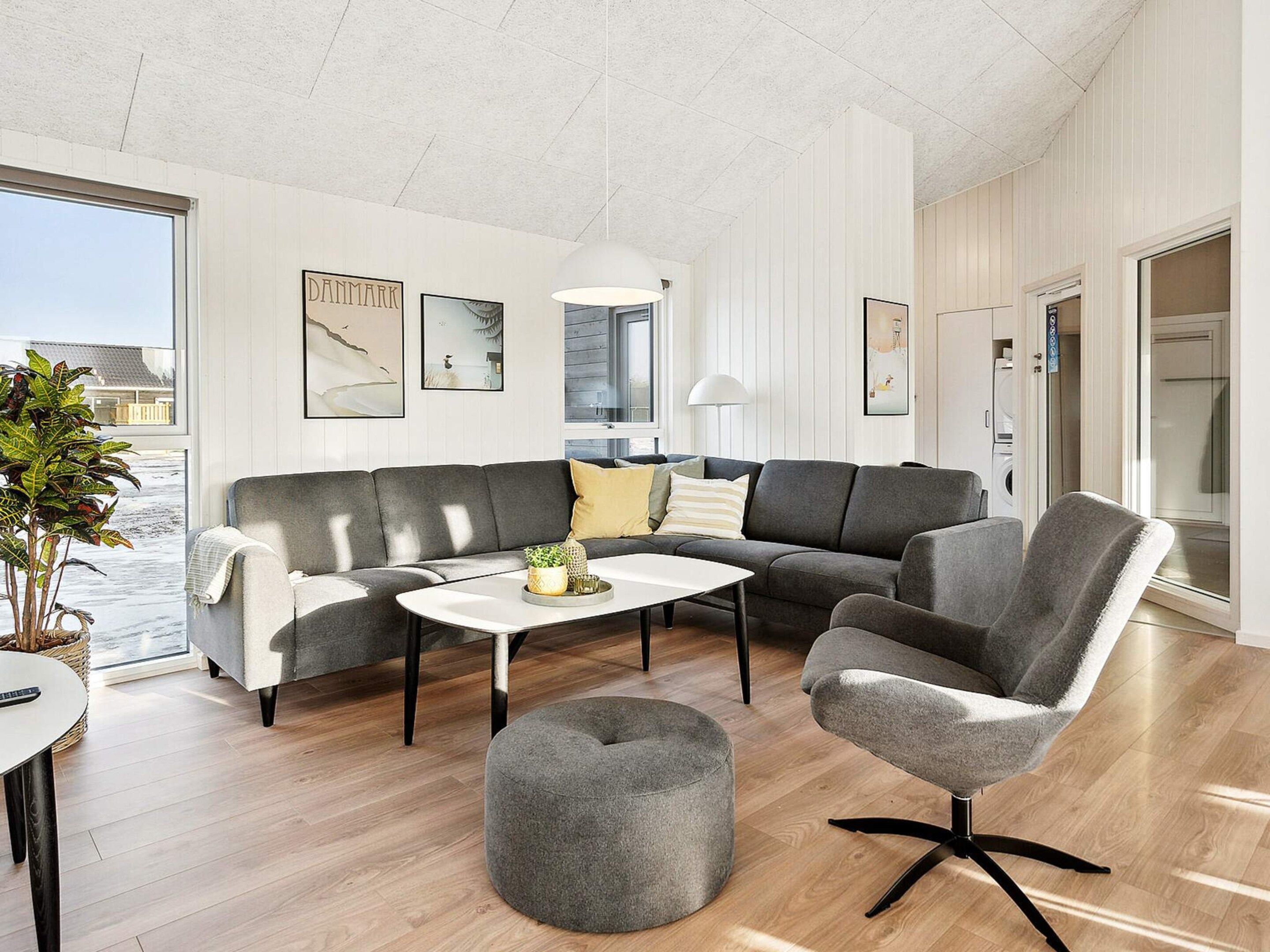 A modern living room with a grey sofa, a round ottoman, and a grey armchair. There are framed pictures on the wall, a white coffee table, and a large window providing a view outside.