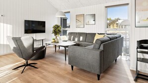 Un salon moderne avec un canapé gris, un fauteuil noir et une table basse. Un téléviseur à écran plat est fixé au mur, ainsi qu’une plante en pot et des œuvres d’art.