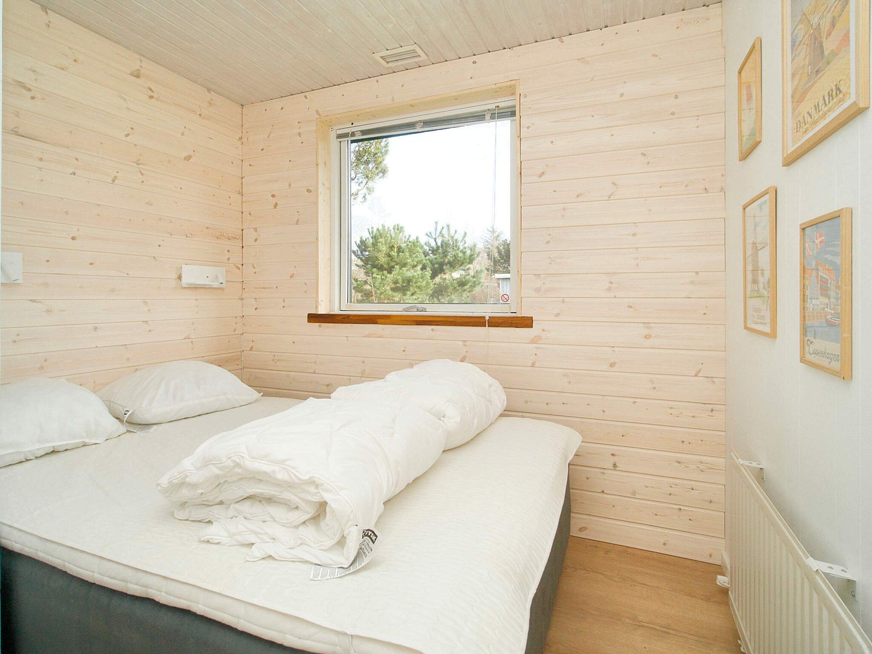 Ein Zimmer mit Holzwänden, einem Bett mit weißer Bettwäsche, einem Fenster mit Blick auf Bäume und einer Wand mit gerahmten Bildern.