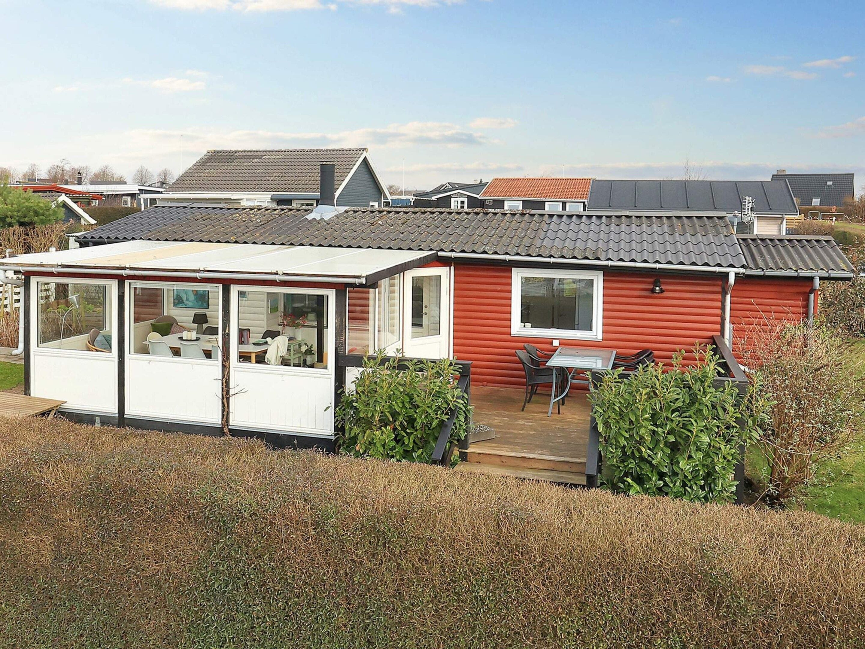 A red mobile home with a covered patio area, a small deck, and a window overlooking a grassy area.