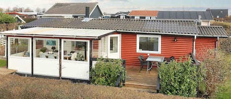A red mobile home with a covered patio area, a small deck, and a window overlooking a grassy area.