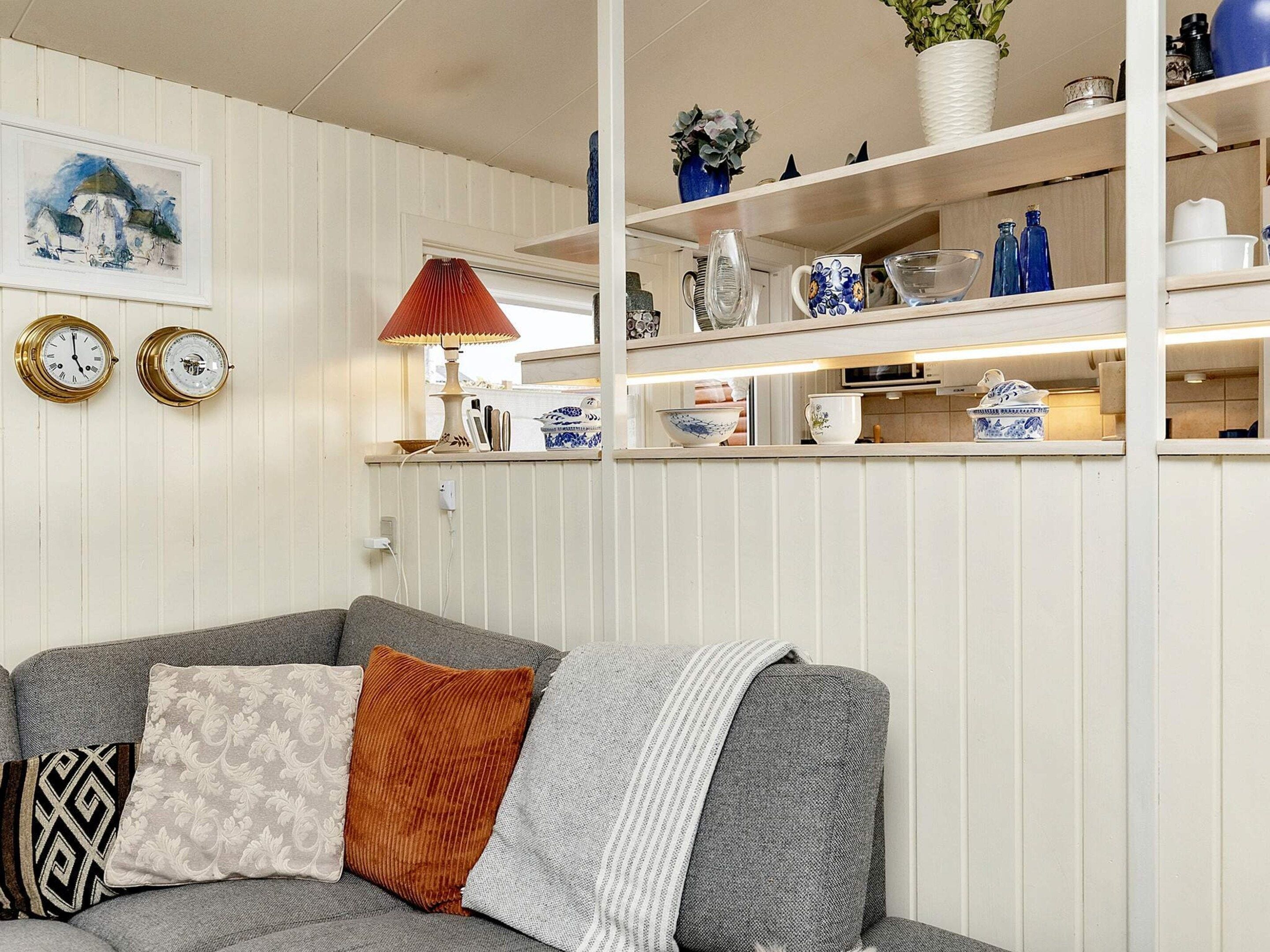 A living room with a grey sofa, patterned cushions, a red lamp, and a white wall with a framed picture and two clocks.