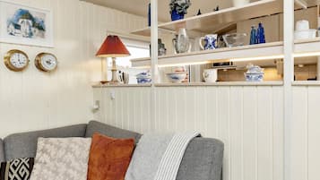 A living room with a grey sofa, patterned cushions, a red lamp, and a white wall with a framed picture and two clocks.