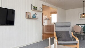 Una sala de estar moderna con un televisor de pantalla plana, un sillón gris, una zona de comedor con una mesa y sillas, y una cocina al fondo.