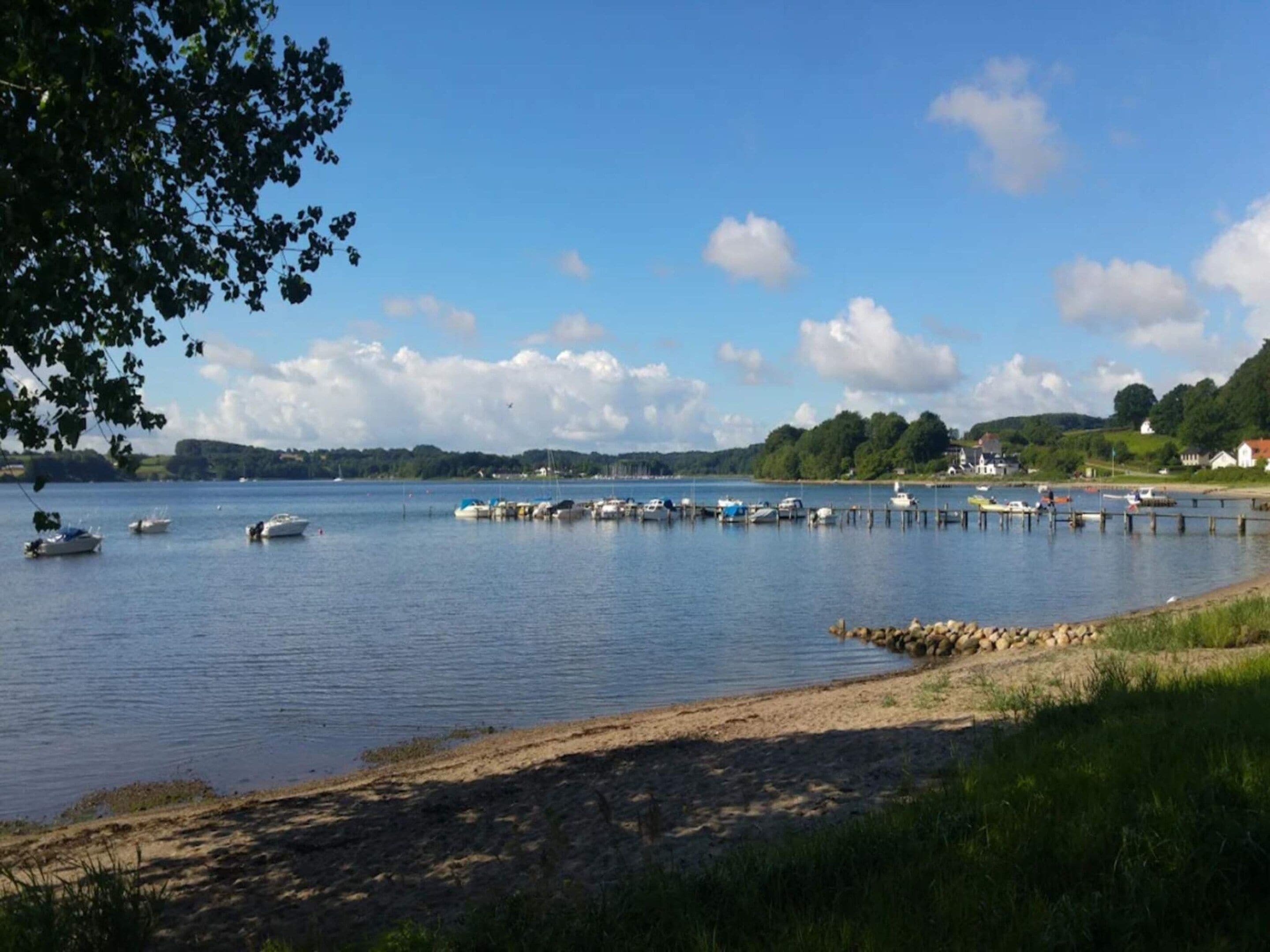 A calm lake with boats docked at a pier, surrounded by greenery and houses.