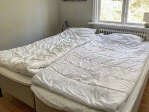 Cama de casal com roupa de cama branca num quarto com janela e radiador.