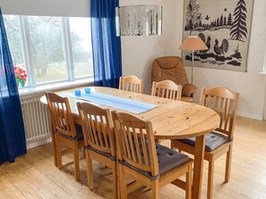 A dining area with a wooden table and chairs, a window with blue curtains, and a painting on the wall. - 8 Person Holiday Home in Anneby (Aneby)