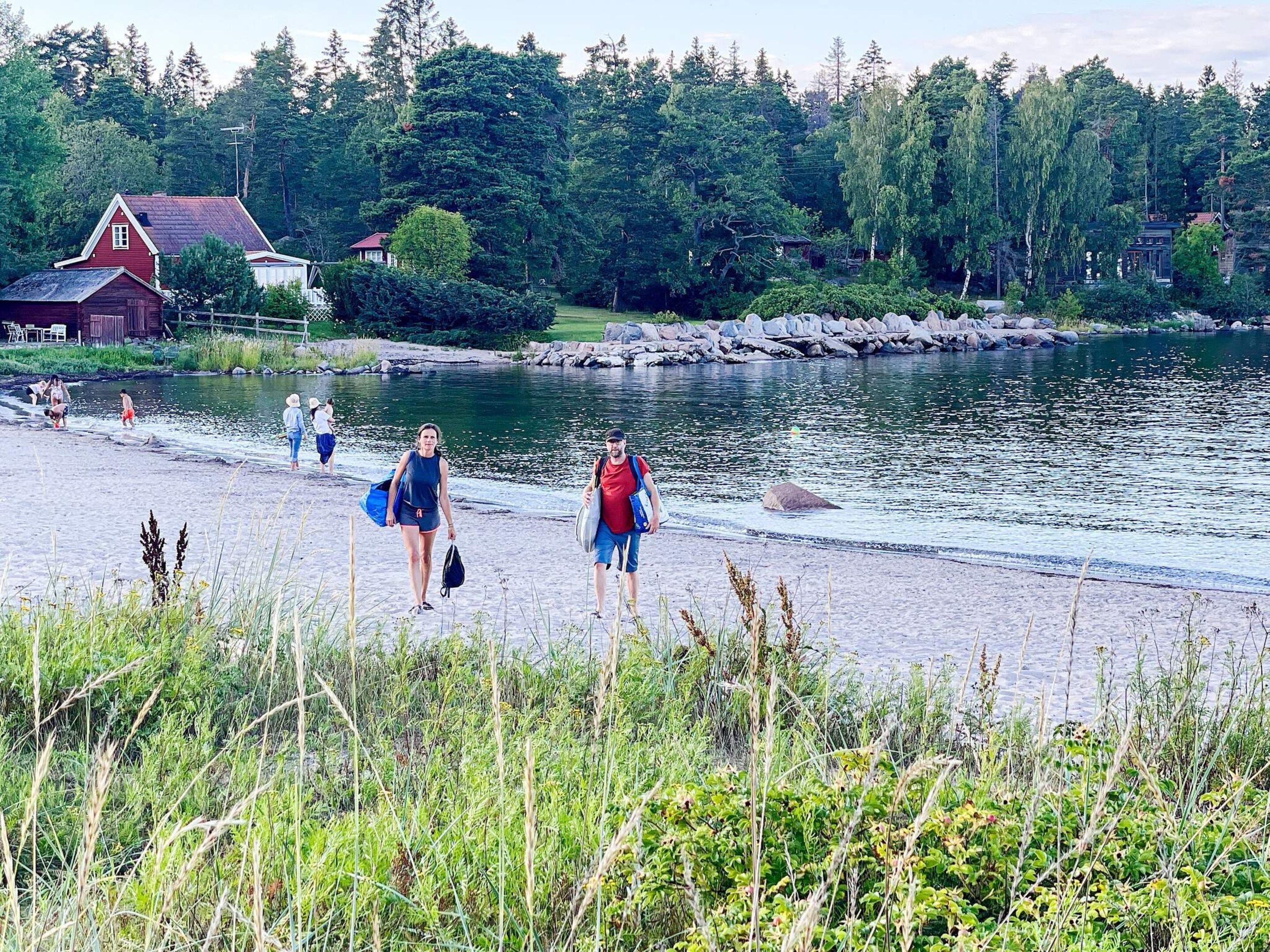 A lakeside walkway with people walking, surrounded by greenery and a house in the background.