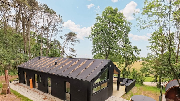 A modern black cabin with large windows, a patio, and a hot tub in a wooded area. - 5 Star Holiday Home in Frederikshavn (Frederikshavn)