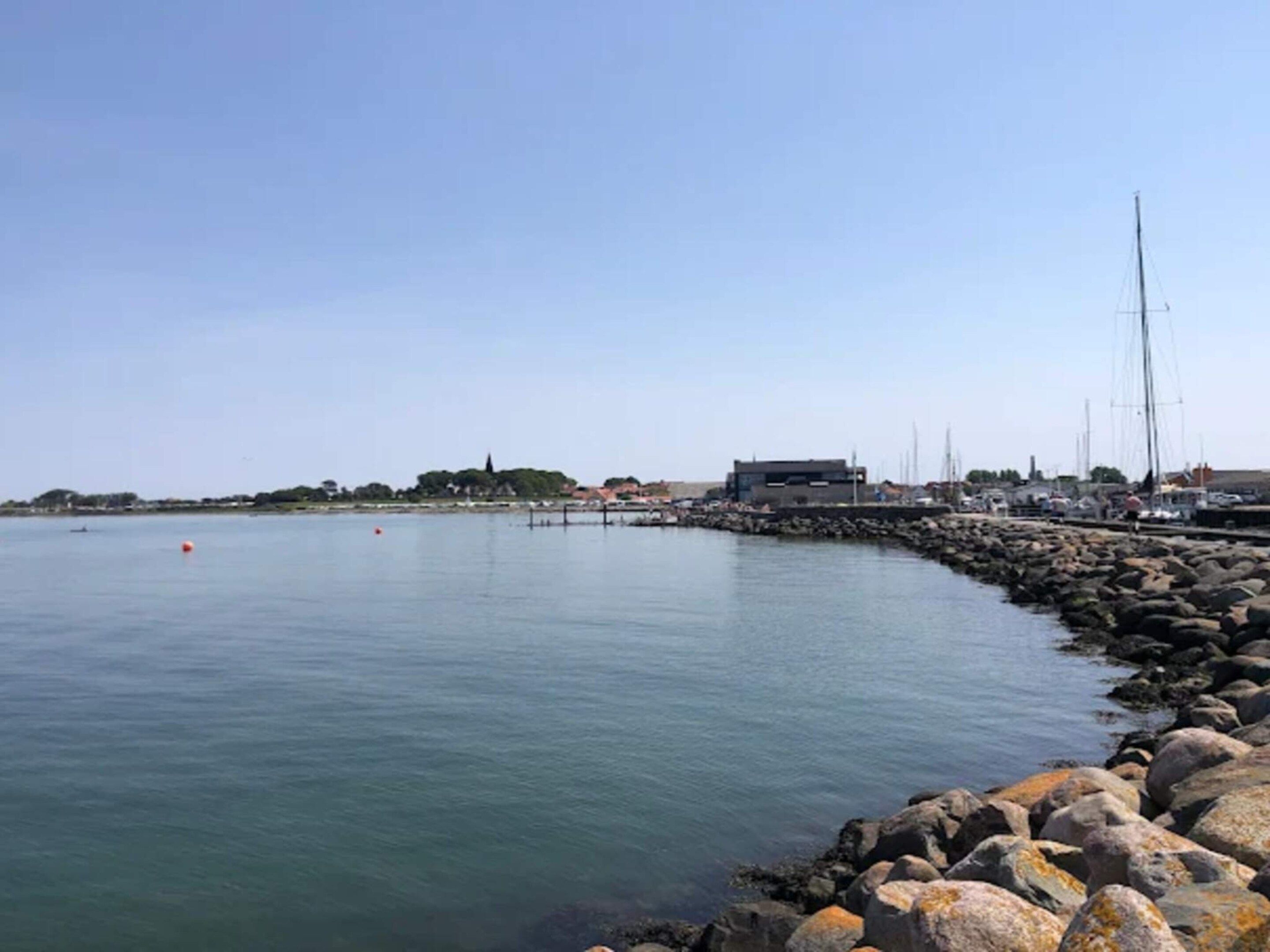 A coastal marina with boats, a rocky breakwater, and buildings in the background.