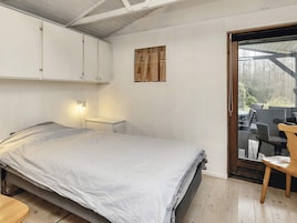 Un dormitorio con cama, mesita de noche, lámpara de pared, una silla de madera y un ventanal corredero que da a un espacio exterior con mobiliario.