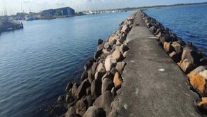 Ein Betonweg führt zu einer felsigen Küstenlinie mit ruhigem Wasser, einem entfernten Gebäude und einem klaren Himmel.